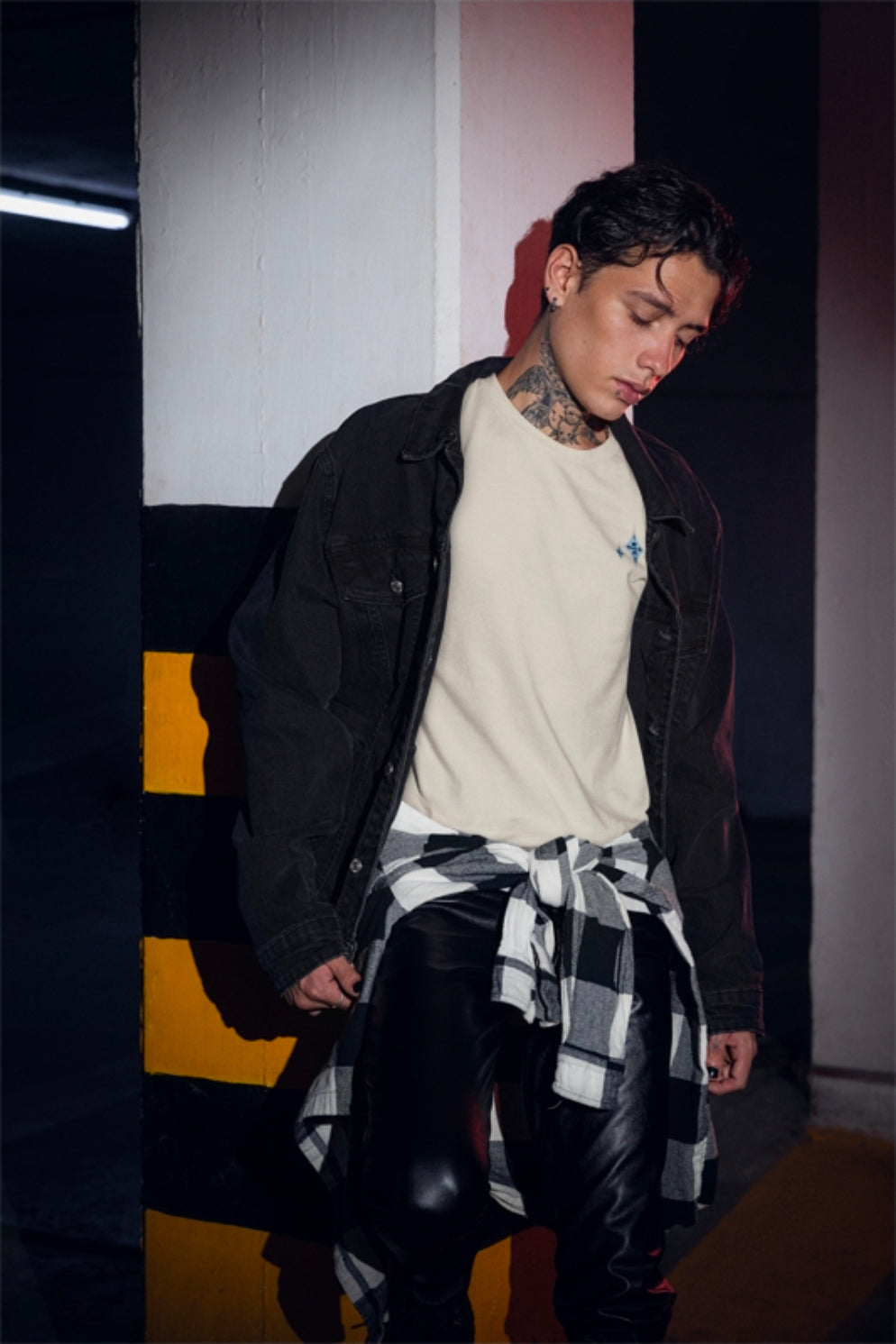 A person wearing a black denim jacket over a cream t-shirt, with a black and white plaid shirt tied around his waist and black leather pants, standing in a dimly lit modern hallway.
